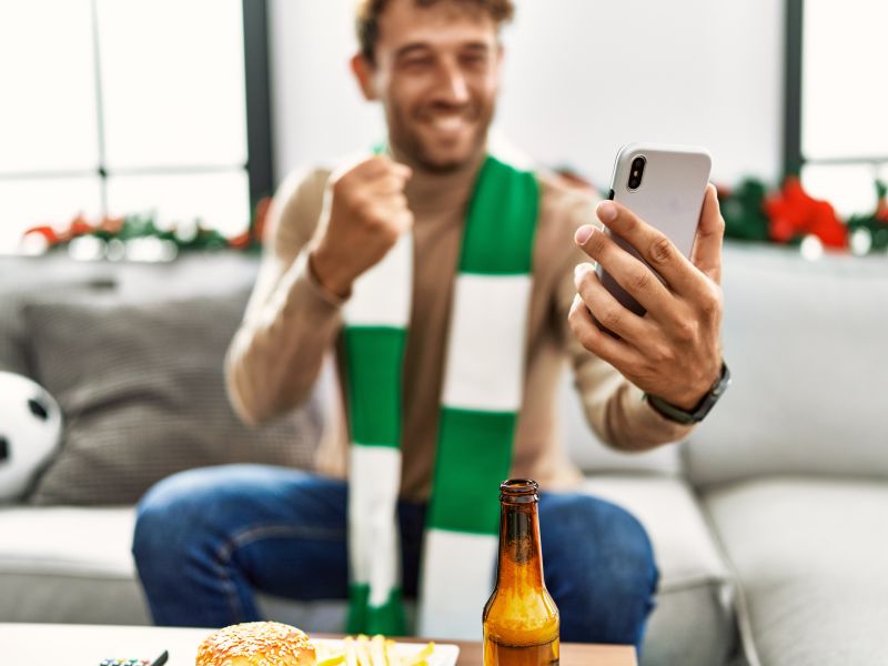 Young hispanic man supporting soccer match using smartphone sitting by christmas decor home